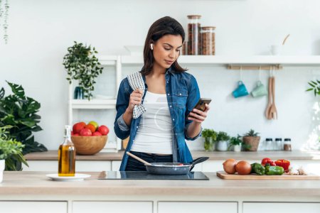 Téléchargez les photos : Tourné de belle femme souriante cuisine tout en utilisant le téléphone portable dans la cuisine à la maison - en image libre de droit