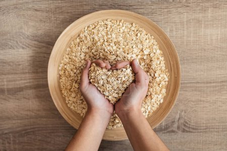 Photo for Close up of woman holding heart-shaped hands of oatmeal from a wooden bowl in the kitchen at home - Royalty Free Image