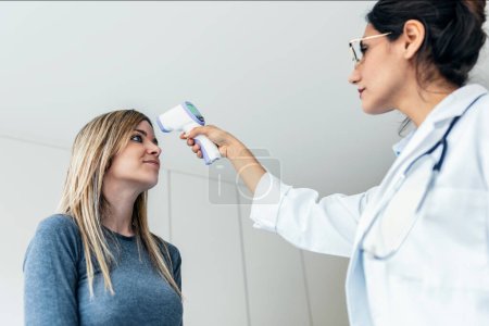Foto de Tiro de alegre hermosa doctora tomando temperatura de paciente mujer con termómetro en consulta médica - Imagen libre de derechos