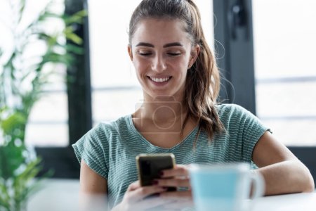 Téléchargez les photos : Prise de vue d'une jeune femme d'affaires concentrée utilisant son téléphone portable tout en travaillant avec un ordinateur portable dans la cuisine à la maison - en image libre de droit
