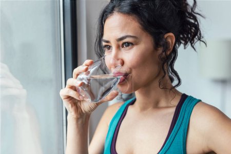 Photo for Shot of beautiful sporty woman drinking a glass of water while standing near the window at home - Royalty Free Image