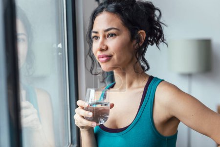 Aufnahme einer schönen sportlichen Frau, die ein Glas Wasser trinkt, während sie zu Hause am Fenster steht
