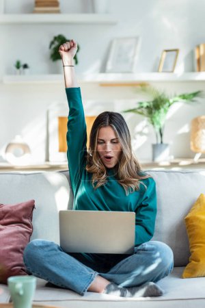 Photo for Shot of pretty young woman celebrating something while using her laptop on the sofa at home. - Royalty Free Image