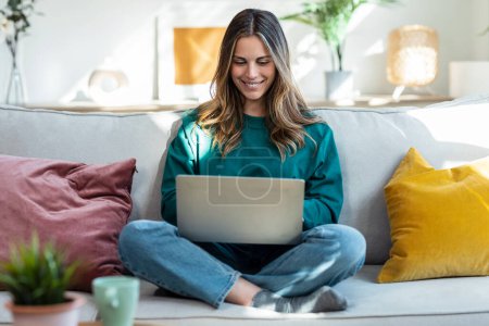 Téléchargez les photos : Prise de vue de belle femme aimable travaillant avec un ordinateur portable tout en étant assis sur le canapé dans le salon à la maison - en image libre de droit