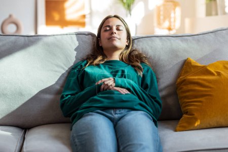 Photo for Shot of young tired woman relaxing on couch with pillow for stomachache in living room at home. - Royalty Free Image