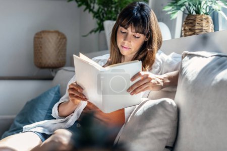 Aufnahme einer hübschen jungen Frau beim Lesen eines Buches, während sie zu Hause auf dem Sofa sitzt.
