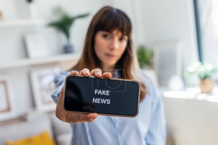 Téléchargez les photos : Prise de vue d'une femme sérieuse tenant le téléphone portable avec le message fausses nouvelles à la maison - en image libre de droit