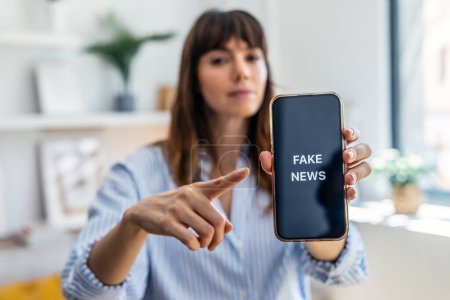 Photo for Shot of serious woman holding the mobile phone with the message fake news at home - Royalty Free Image