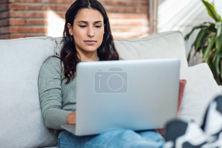 Foto de Foto de una hermosa mujer joven que trabaja con su computadora portátil mientras está sentada en un sofá en casa. - Imagen libre de derechos