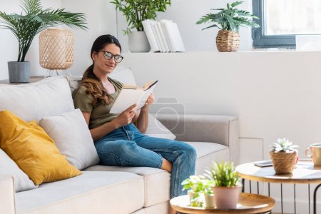 Aufnahme einer selbstbewussten Frau beim Lesen eines Buches, während sie zu Hause auf dem Sofa sitzt.