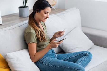Foto de Tiro de mujer segura de pagar algo en línea con su tarjeta de crédito en el ordenador portátil mientras está sentado en el sofá en casa. - Imagen libre de derechos