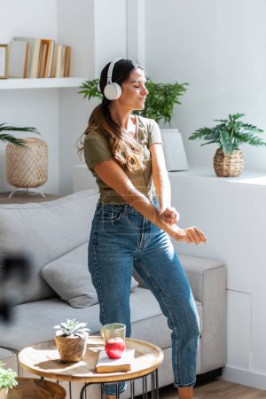 Foto de Foto de mujer feliz motivada cantando y bailando en el sofá mientras escucha música con auriculares en casa. - Imagen libre de derechos