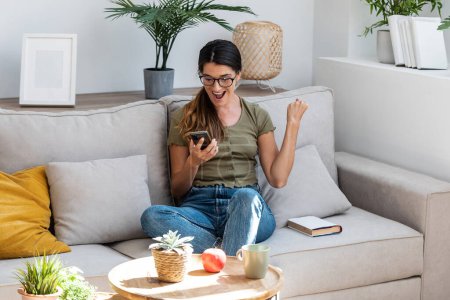 Téléchargez les photos : Prise de vue de jeune femme excitée célébrant quelque chose tout en utilisant smartphone détente dans le canapé à la maison. - en image libre de droit