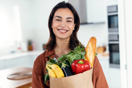 Téléchargez les photos : Portrait de belle jeune femme tenant un sac en papier plein de légumes tout en regardant la caméra dans la cuisine à la maison - en image libre de droit