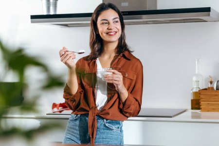 Photo for Shot of beautiful happy woman eating a yoghurt while looking forwards in the kitchen at home - Royalty Free Image