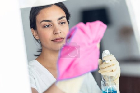 Téléchargez les photos : Potrait de belle jeune femme nettoyant le miroir avec un chiffon rose et pulvérisation à la maison - en image libre de droit