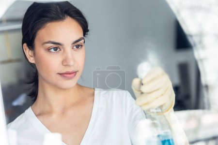 Foto de Potrait de hermosa joven limpiando el espejo con un spray en casa - Imagen libre de derechos