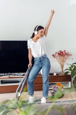 Téléchargez les photos : Prise de vue d'une jeune femme heureuse écoutant et dansant de la musique tout en nettoyant le sol du salon avec un nettoyant vaccinal - en image libre de droit