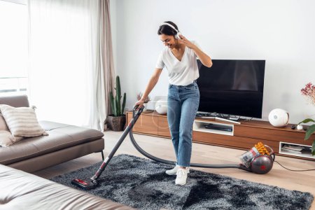 Photo for Shot of young happy woman listening to music while cleaning the living room floor with a vaccum cleaner - Royalty Free Image
