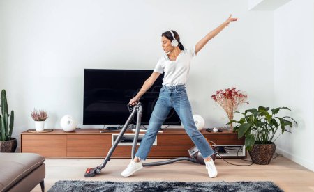 Foto de Tiro de mujer feliz joven escuchando y bailando música mientras se limpia el piso de la sala de estar con un limpiador de vaccum - Imagen libre de derechos
