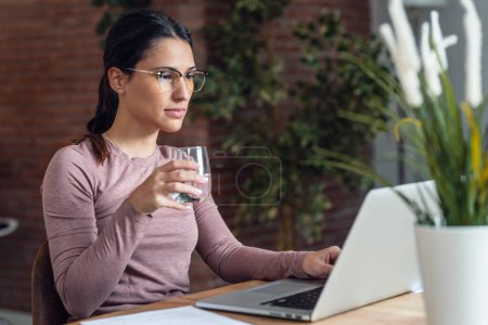 Téléchargez les photos : Prise de vue de belle jeune femme travaillant avec PC tout en buvant un verre d'eau - en image libre de droit