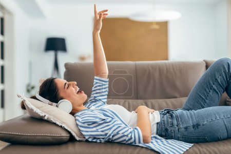 Foto de Foto de hermosa mujer cantando mientras escucha música tumbada en el sofá en la sala de estar en casa - Imagen libre de derechos