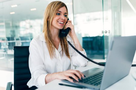 Foto de Tiro de elegante atractiva mujer de negocios hablando con el teléfono mientras trabaja con el ordenador portátil en una startup moderna - Imagen libre de derechos