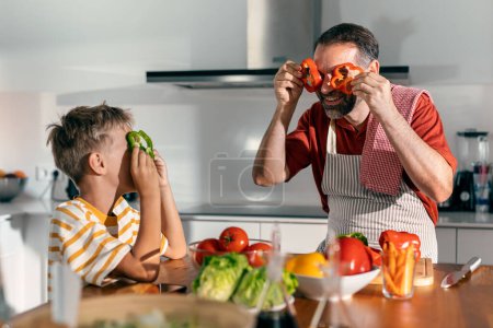 Photo for Shot of funny father and son having fun and playing with peppers slices on the eyes in the kitchen at home - Royalty Free Image