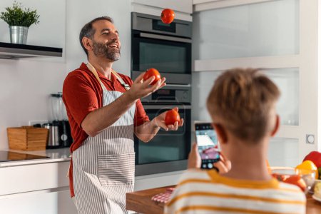 Foto de Tiro de divertido padre haciendo malabares con tomates mientras su hijo lo graba con un teléfono móvil en la cocina - Imagen libre de derechos
