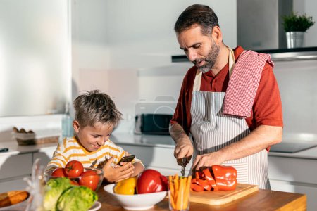 Foto de Un disparo de padre guapo enseñando a su hijo a cocinar mientras el hijo lo graba con el teléfono móvil en la cocina en casa - Imagen libre de derechos