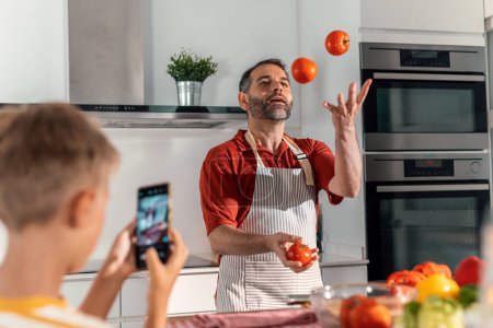 Photo for Shot of funny father juggling tomatoes while his son is recording him with a mobile phone in the kitchen - Royalty Free Image