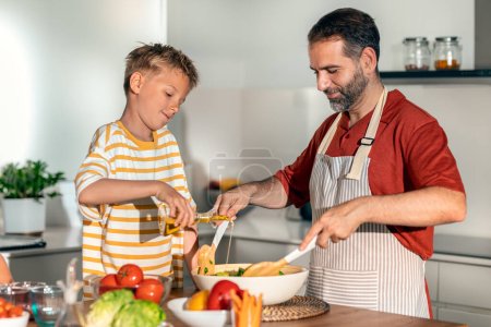 Foto de Foto de padre guapo cocinando ensalada saludable con su hijo en la cocina en casa - Imagen libre de derechos