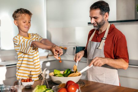 Téléchargez les photos : Tourné de beau père cuisine salade saine avec son fils dans la cuisine à la maison - en image libre de droit