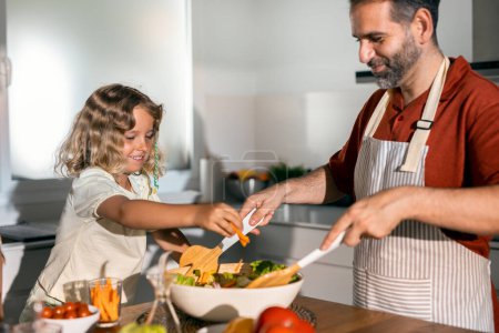 Foto de Foto de padre guapo cocinando ensalada saludable con su amable hija en la cocina en casa - Imagen libre de derechos