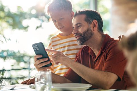 Téléchargez les photos : Beau père et son fils mignon en utilisant smartphone avant le déjeuner dans la cuisine à la maison - en image libre de droit