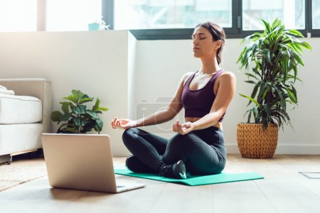 Foto de Tiro de mujer joven y deportiva haciendo ejercicios de yoga después de clases de gimnasia en línea a través de computadora portátil en el suelo en casa. - Imagen libre de derechos