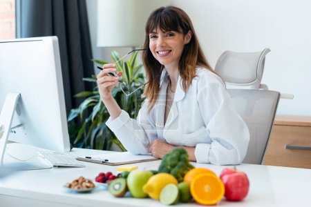 Aufnahme der schönen smarten Ernährungsberaterin, die mit dem Computer arbeitet, während sie in die Kamera in der Ernährungsberatung schaut