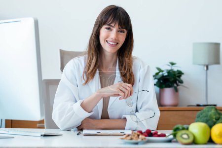 Foto de hermosa mujer nutricionista inteligente que trabaja con el ordenador mientras mira a la cámara en la consulta nutricionista