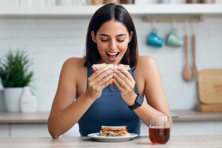 Prise de vue de belle femme sportive mangeant sanwich sain tout en regardant la caméra dans la cuisine à la maison