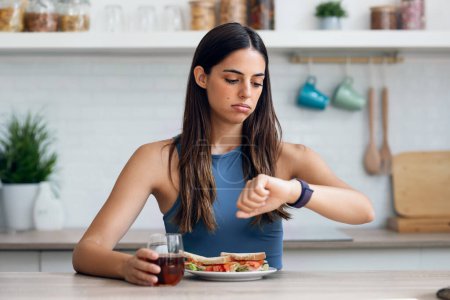 Foto de Tiro de mujer deportiva hambrienta esperando el tiempo para comer mientras mira reloj después de ayunar en la cocina en casa - Imagen libre de derechos