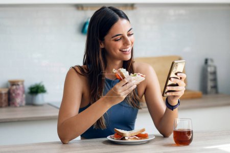 Photo for Shot of beautiful sporty woman eating healthy sanwich while using smartphone in the kitchen at home - Royalty Free Image
