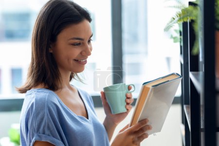 Téléchargez les photos : Tourné de belle jeune femme buvant une tasse de café tout en cherchant un livre dans la librairie à la maison - en image libre de droit