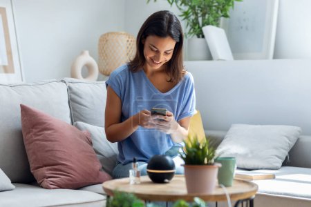 Photo for Shot of beautiful woman using smartphone while essential oil aroma diffuser humidifier the air in living room at home - Royalty Free Image