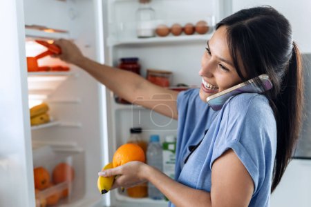 Foto de Dispara a una hermosa mujer joven tomando algo de comida saludable de la nevera mientras habla con el teléfono inteligente en la cocina en casa - Imagen libre de derechos