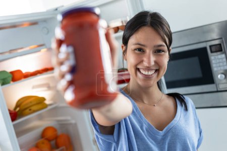 Foto de Foto de hermosa mujer sonriente sosteniendo tarro de tomate mientras mira la cámara frontal de la nevera en la cocina. - Imagen libre de derechos