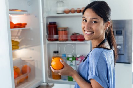 Photo for Shoot of beautiful young woman taking an orange from the fridge while looking at camera in the kitchen at home - Royalty Free Image