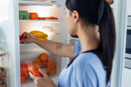 Téléchargez les photos : Prise de vue de belle jeune femme hésitant à manger devant le réfrigérateur dans la cuisine. - en image libre de droit