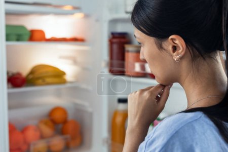 Aufnahme einer selbstbewussten jungen Frau, die zögert, vor dem Kühlschrank in der Küche zu essen.