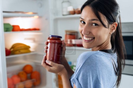 Téléchargez les photos : Tourné de belle femme tenant pot de tomate tout en regardant la caméra devant le réfrigérateur dans la cuisine. - en image libre de droit