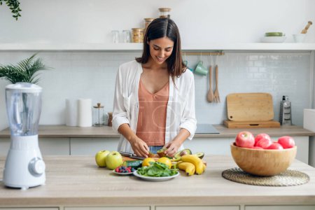 Téléchargez les photos : Tourné de femme souriante coupant pepino pour préparer un jus de désintoxication dans la cuisine à la maison. - en image libre de droit